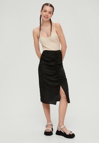Beige geribd tanktop met knopen, gecombineerd met een zwarte, midi-rok met zijruching en een hoge split, en platformsandalen.