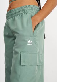 Grüne Cargo-Hosen mit einem elastischen Bund, Seitentaschen und einem weißen Adidas-Logo. Glattes Material mit einer lockeren Passform.