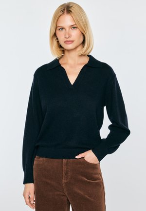 Blondynka w czarnym swetrze z długim rękawem i dekoltem w serek oraz brązowych spodniach sztruksowych stoi z jedną ręką w kieszeni.