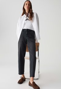Jeans neri a vita alta con tasche anteriori, abbinati a una camicia bianca con bottoni e a una sciarpa a fantasia. Mocassini marroni completano l'outfit.