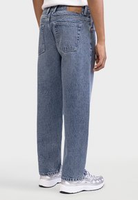 Ανοιχτό μπλε τζιν με άνετη εφαρμογή, με δερμάτινο ταμπελάκι στη μέση πίσω και δύο πίσω τσέπες. Φορεμένο με λευκά και γκρι αθλητικά παπούτσια.