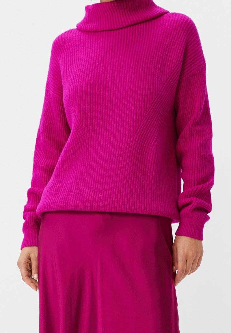 Femme portant un pull à col roulé côtelé rose vif et une jupe rose assortie, debout les mains détendues le long du corps.