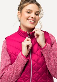 Fuchsia gesteppte Weste mit Stehkragen und einem Frontreißverschluss, kombiniert mit einem strukturierten pinken Langarmoberteil mit kontrastierenden Ärmeln.