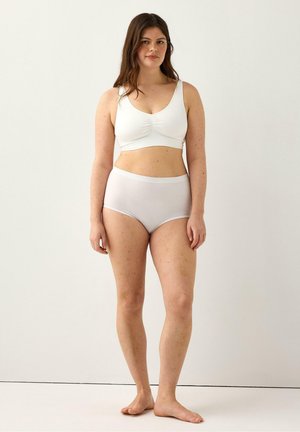 Kvinde med langt brunt hår iført en hvid bh og højttaljede trusser, stående barfodet mod en ensfarvet lys baggrund.