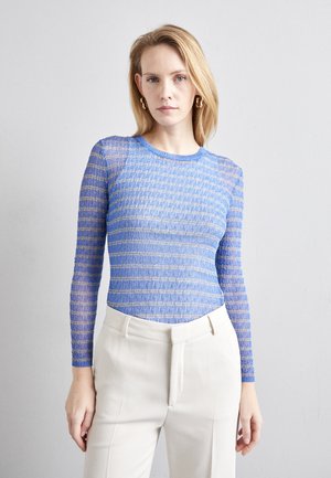 Blue long-sleeve top with a textured pattern of horizontal stripes in varying shades of blue and subtle gold accents, paired with cream trousers.