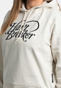 Crèmekleurige hoodie met een trekkoord. Voorzien van vetgedrukt zwart lettertype "Hant Breiker" over de borst. Gemaakt van zachte stof met ribbels aan de uiteinden.