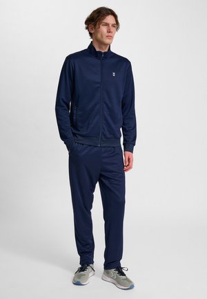 Marineblauwe trainingspak gemaakt van soepel materiaal, met een hoge kraag en een ritssluiting aan de voorkant. De broek heeft een elastische tailleband. Grijze sneakers maken de look af.