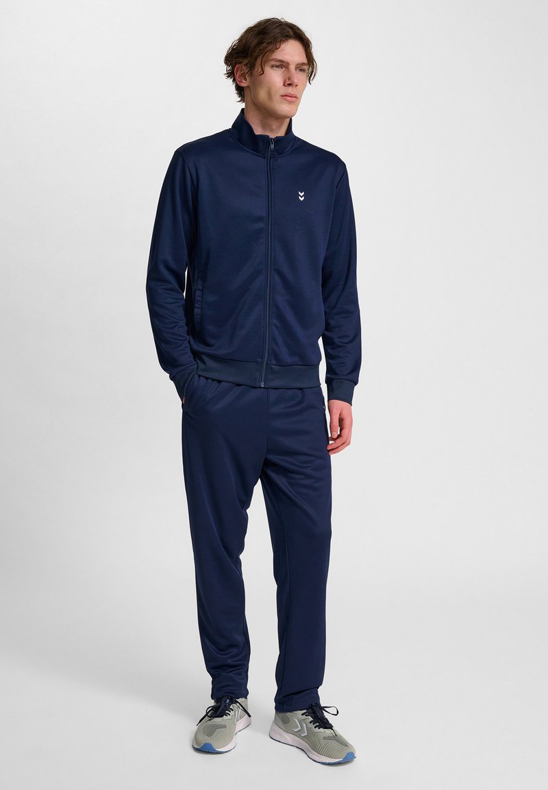 Tuta da ginnastica blu navy realizzata in tessuto morbido, con colletto alto e zip frontale. I pantaloni hanno una vita elastica. Sneakers grigie completano il look.