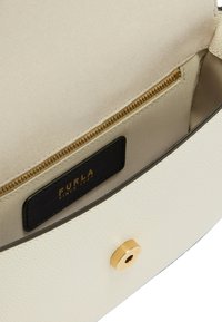Interno di una borsa in pelle crema con fodera beige, tasca con zip dorata e etichetta nera con la scritta "FURLA" in oro. Dettaglio in oro a forma circolare.