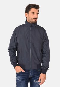 Giacca bomber blu navy con colletto alto, zip frontale e polsini a costine. Presenta tasche laterali e un tessuto liscio e leggero.