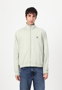 Lys grønn zip-up jakke med høyt krage, elastiske mansjetter og kant; har et lite logo-patch på venstre bryst og sidelommer.