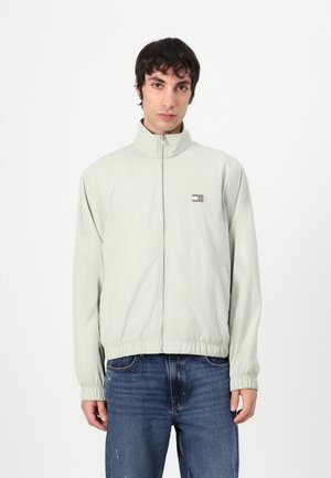 Light green zip-up jacket with a high collar, elastic cuffs, and hem; features a small logo patch on the left chest and side pockets.
