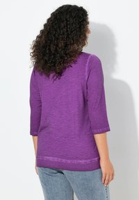 Fioletowa bluzka z rękawami do łokcia, wykonana z teksturowanej tkaniny. Brzeg dolny ozdobiony przeszyciami, zestawiona z jasnoniebieskimi dżinsami.