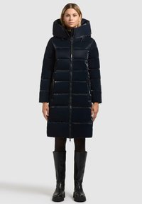 Piumino blu navy con grande cappuccio, design a righe orizzontali, zip frontale e tasche laterali. Indossato sopra un completo nero e stivali sopra il ginocchio.