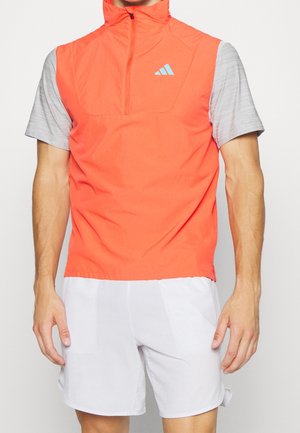 Mann trägt eine leuchtend orange ärmellose Weste mit Reißverschluss über einem hellgrauen Kurzarmshirt und weißen Sportshorts vor einfarbigem Hintergrund.