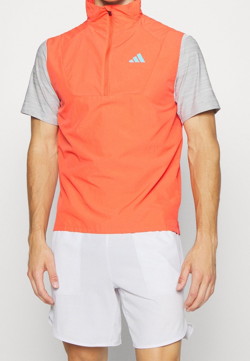 Homme portant un gilet sans manches zippé orange vif sur un t-shirt gris clair à manches courtes et un short de sport blanc sur fond uni.