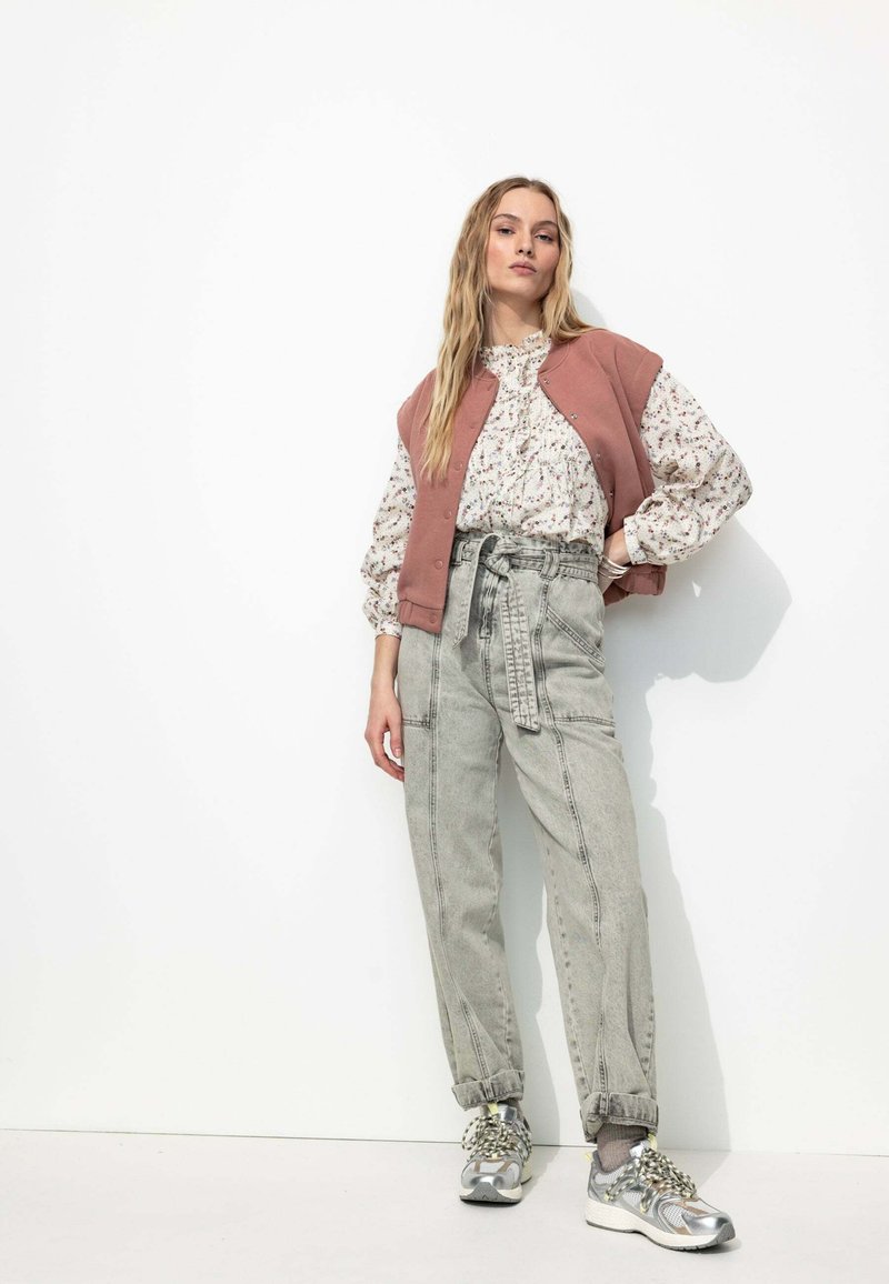 Débardeur de sport rose sur une blouse à fleurs, associé à un jean gris taille haute et des baskets argentées. Présente une coupe ample et des poignets froncés.