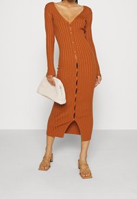 Robe en tricot côtelé couleur rouille avec manches longues, col en V et fermeture à boutons sur le devant. Longueur mi-mollet avec une fente à l'ourlet. Sac à main tissé beige.