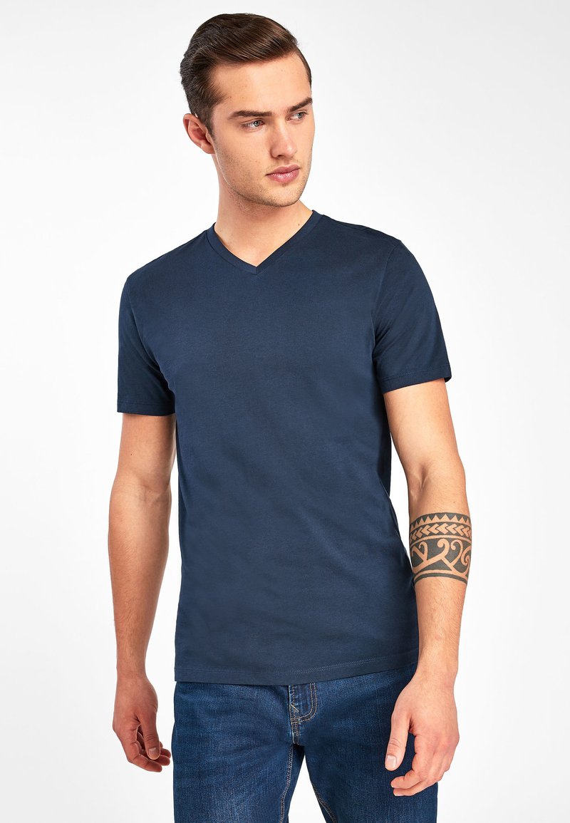 Temno modra majica s potiskom v obliki črke V, izdelana iz mehkega materiala, z kratkimi rokavi in prilegajočim krojem, v kombinaciji z jeans hlačami.