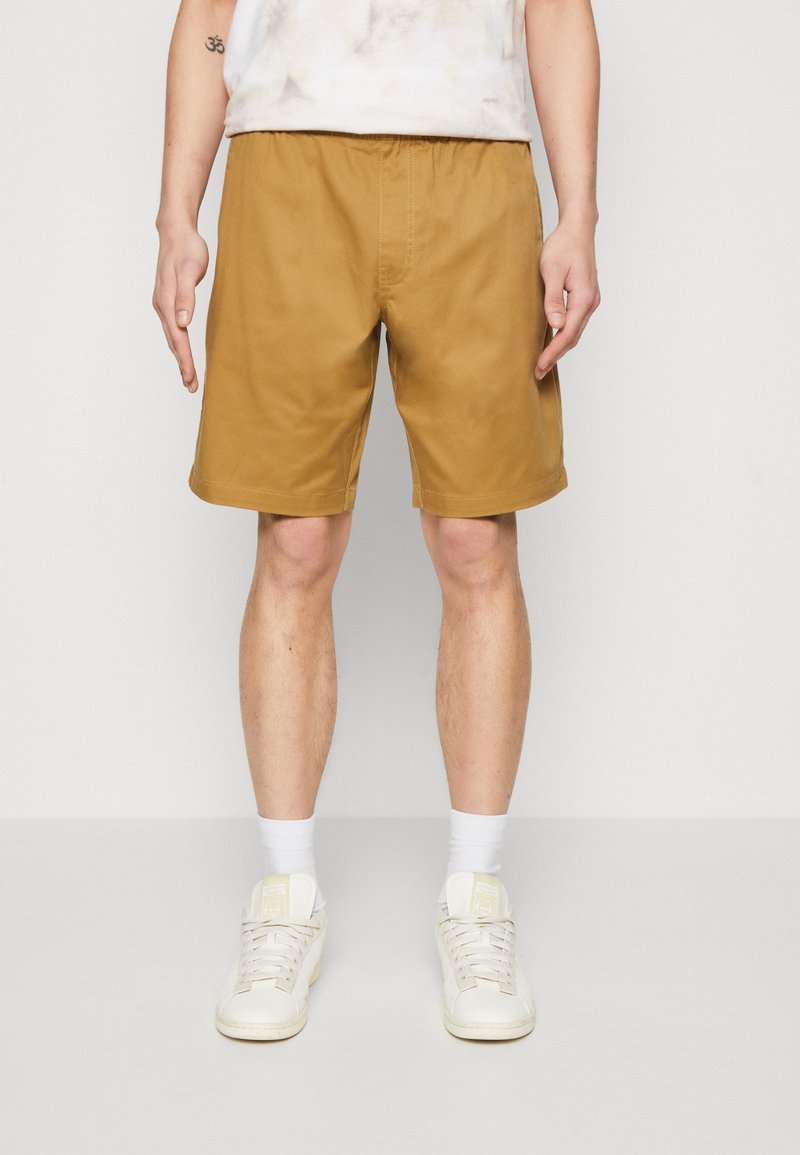 Homme portant un short jaune moutarde, des baskets blanches, des chaussettes blanches et un haut de couleur claire, debout devant un fond uni.