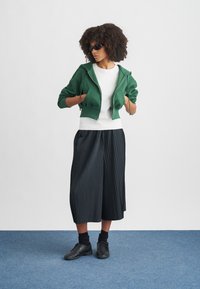 Zelená skrátená mikina s kapucňou, biele tričko, čierne plisované široké nohavice, čierne tenisky a čierne slnečné okuliare. Hladké textúry, neformálny dizajn.