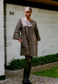 Leopardenmuster, knielanges Kleid mit Dreiviertelärmeln und Seitentaschen. Aus strukturiertem Stoff gefertigt. Schwarze Strumpfhosen und schlichte schwarze Schuhe.