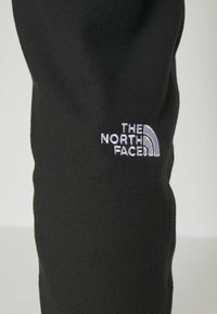 Svart fleece-tyg med en slät yta; har en vit broderad logotyp "THE NORTH FACE" och en halvcirkelformad designaccent.