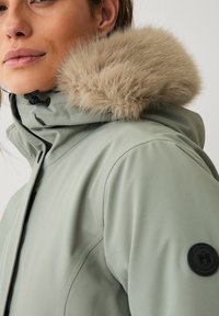 Lichtgroene waterdichte jas met een nepbont capuchon, voorzien van een zwarte badge op de mouw en een hoge kraag voor extra warmte.