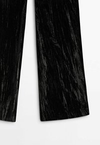 Gros plan sur un pantalon large en velours noir écrasé sur fond blanc, mettant en valeur la texture et la brillance du tissu.