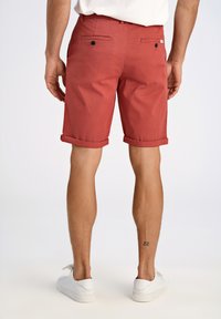 Shorts en coton rouge avec des revers, dotés de deux poches arrière avec des boutons noirs. Modèle portant des baskets blanches.