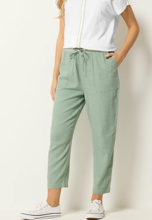 Frau trägt salbeigrüne Kordelzughose, weiße Freizeitsneaker und ein weißes Kurzarmhemd mit Stickerei, steht vor einem schlichten Hintergrund.