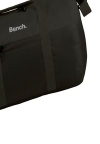 Duffeltas in donkergrijze stof met een gestructureerd oppervlak, voorzien van hexagonale patronen, verstelbare banden en een logo aan de zijkant.