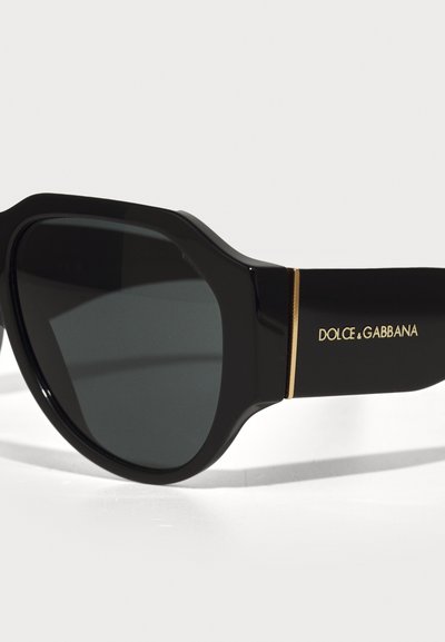 Black sunglasses with a thick frame and dark lenses. Features gold branding on the side and a smooth, polished texture.