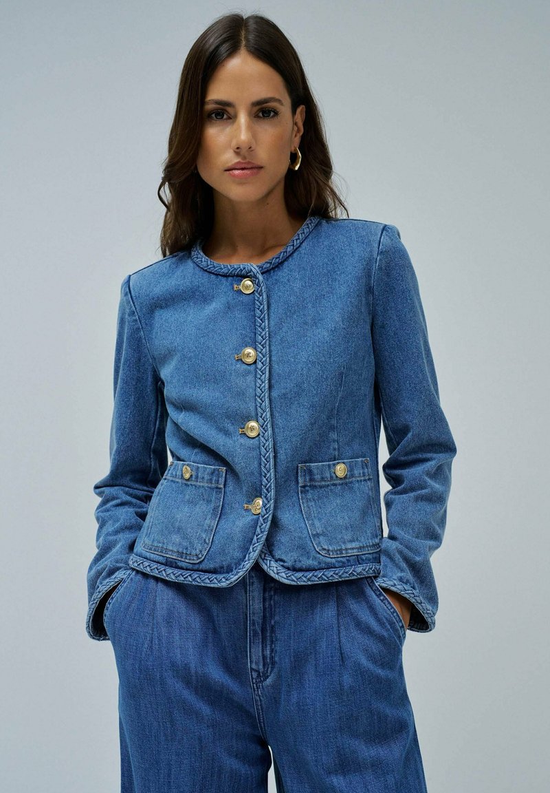 Veste en denim avec fermeture à boutons, garniture tressée et deux poches avant. Couleur bleu clair, tissu légèrement texturé et coupe ajustée.