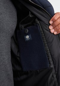 Schwarze Jackeninnenseite mit einer marineblauen Fleece-Tasche mit Knopfverschluss und einem Etikett mit der Aufschrift "Parks Industries". Glatte, matte Textur.