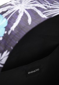 Bolso negro con un patrón floral que presenta flores azules y hojas de palma blancas. El interior es de tela negra, con una etiqueta que dice "even&odd".