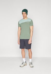 Maglietta verde con pannello diagonale azzurro chiaro, abbinata a pantaloni corti scuri. Indossata con un berretto verde e scarpe da ginnastica beige.