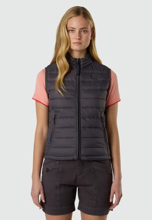 Donna con capelli biondi lunghi indossa un gilet trapuntato nero sopra una maglietta rosa a maniche corte e pantaloncini neri con bottoni, in piedi davanti a uno sfondo semplice.