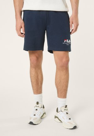 Mężczyzna w granatowych szortach FILA, białych skarpetkach za kostkę i białych butach FILA, stojący na jasnym, jednolitym tle.