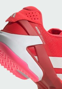 Červená sportovní obuv s texturovaným pleteným svrškem, bílými akcenty a růžovou podešví. Má výrazný design paty a nápis značky.