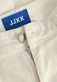 Zbliżenie beżowych spodni z częściowo rozpiętym zamkiem, metalowym guzikiem z grawerem "JJXX" oraz niebieską metką z logo marki.