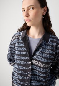 Strikket cardigan med blå og sorte bladmønstre, knaplukninger og en kontrastkrave. Blød tekstur med en afslappet pasform.