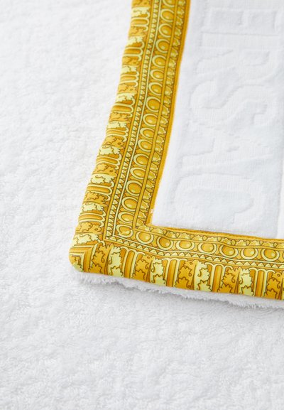Valge froteerätik, millel on pehme tekstuur, kaunistatud kollase ja rohelise mustriga äärisega, milles on ornamentaalsed detailid.