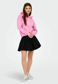 Rosa hoodie med en logotyp framtill, kombinerad med en svart plisserad kjol och vita sneakers. Outfitten har en avslappnad, lager-på-lager-look.