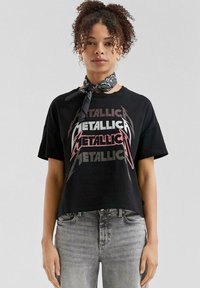 Zwarte cropped t-shirt met het "Metallica" logo in felle kleuren. Model draagt een zwarte bandana om de nek. Zacht, casual stof.