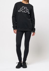 Femme portant un sweat-shirt noir avec un logo blanc de deux silhouettes assises dos à dos, des leggings noirs et des baskets noires, debout sur un fond blanc.