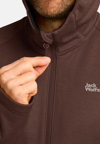 Bruine zip-up jas met een hoge kraag. Kenmerkt zich door een klein logo op de borst, een gladde textuur, en een hand die de ritssluiting vasthoudt.