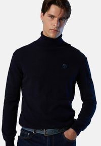 Maglione a collo alto blu navy in tessuto morbido a maglia, con maniche lunghe e un logo discreto sul petto, abbinato a jeans denim blu e una cintura.