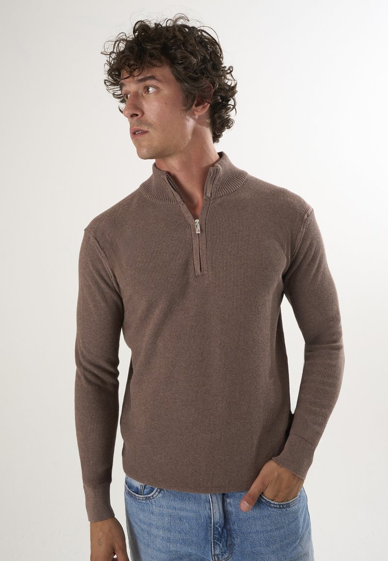 Pull en maille texturée de couleur marron avec fermeture éclair, présentant un col montant et des manches longues. Associé à un jean bleu clair.