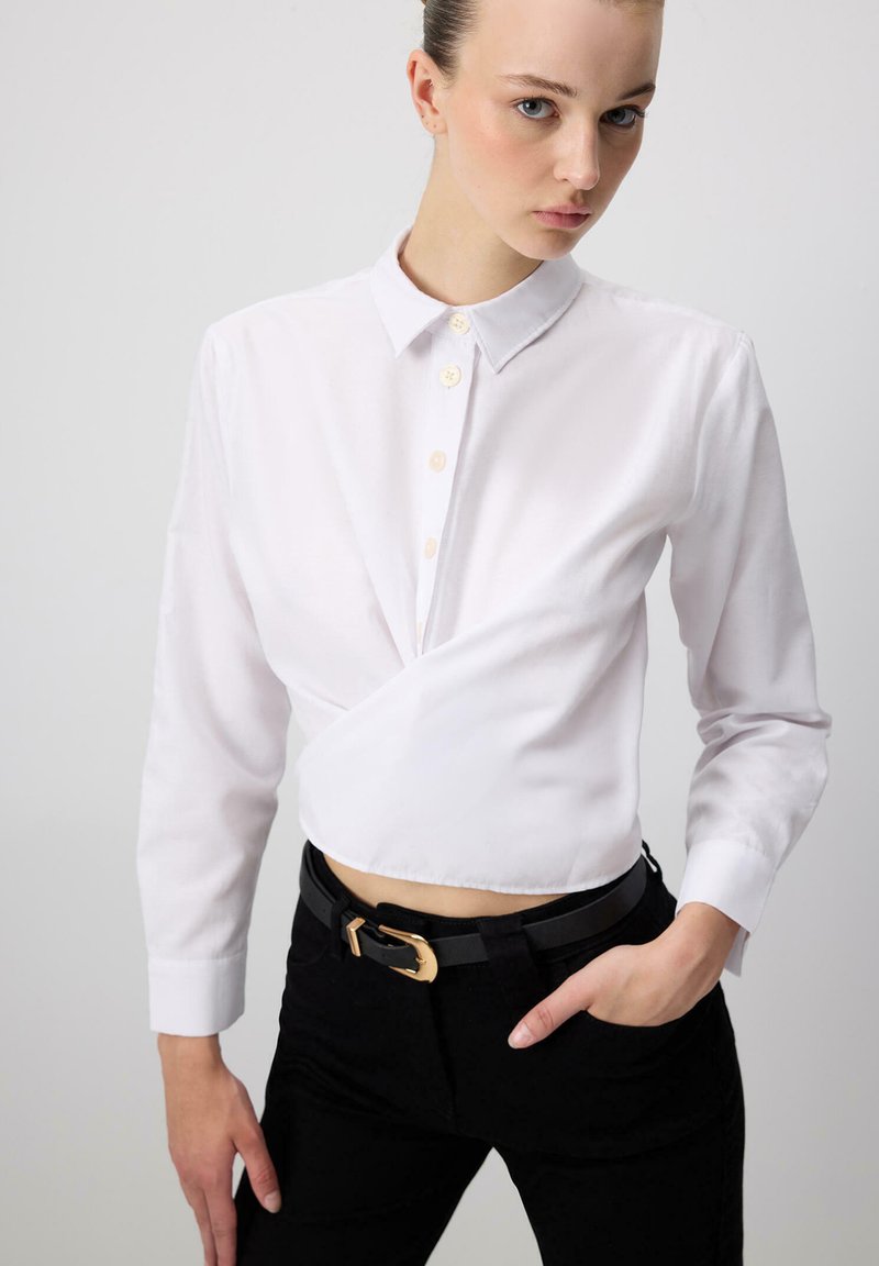 Camicia bianca button-down con un design avvolgente, maniche lunghe e colletto. Abbinata a pantaloni neri e una cintura nera con fibbia dorata.